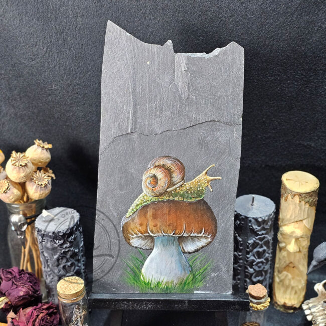 Snail on a toadstool painting on natural Welsh slate, displayed on a black easel with dried botanicals, carved candles
