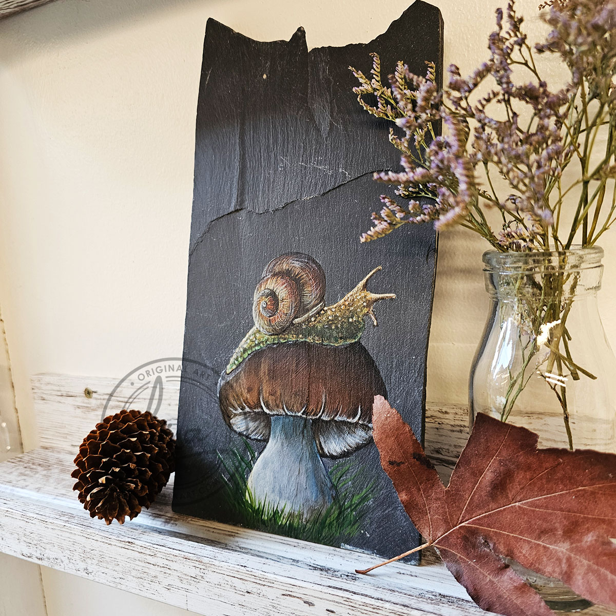 Left angled view of a hand-painted snail on a toadstool on Welsh slate, displayed on a shelf with dark natural décor and dried botanicals.