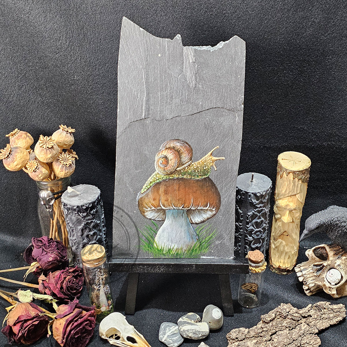 Face-on view of the snail and toadstool slate painting on a black easel, surrounded by dried botanicals and textured carved candles