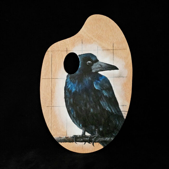 Original painting of a rook on a wooden artist palette by Liisa Clark, featuring iridescent black and blue feathers.