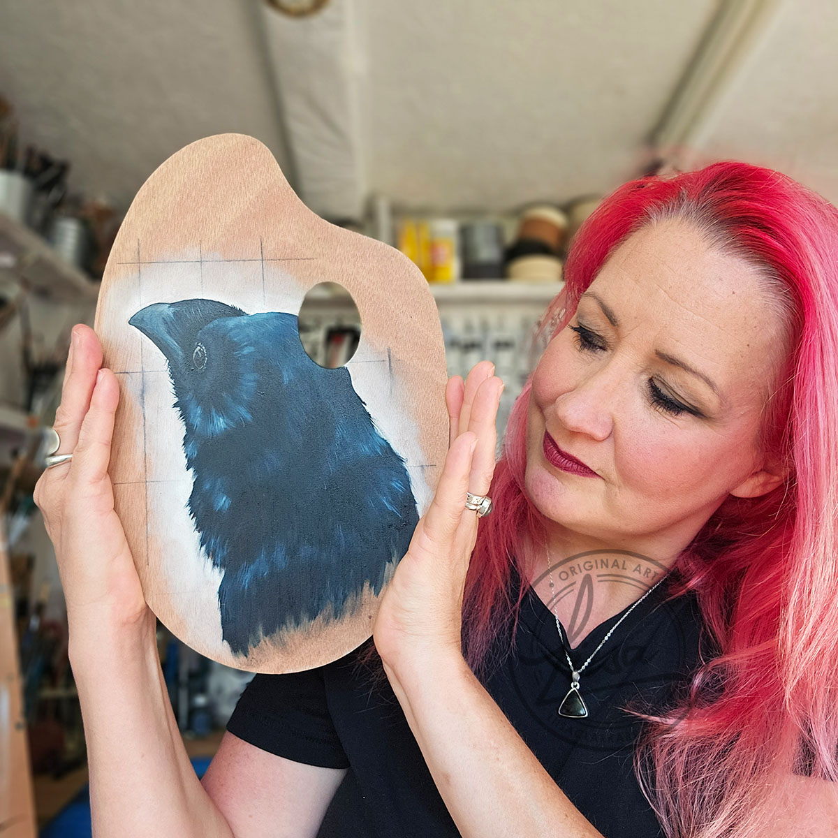 Artist holding crow painting on wooden palette, looking at it.