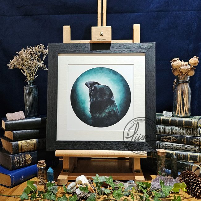 Framed crow artwork titled A Shimmer of Turquoise displayed on a wooden easel surrounded by vintage books, ivy, dried flowers, crystals and natural history styling against a dark backdrop