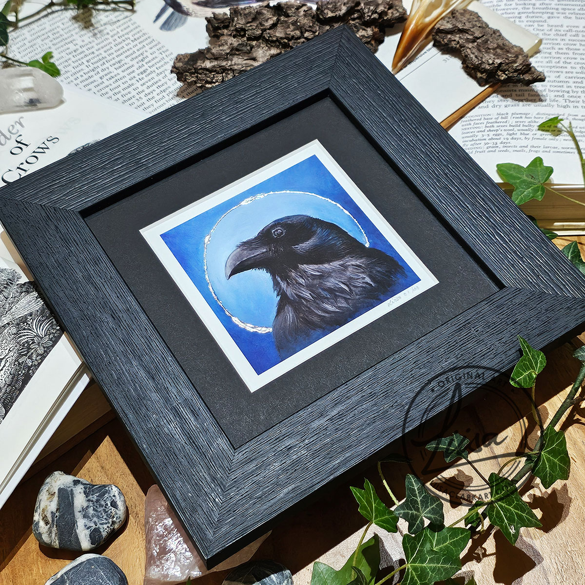 Angled photograph of a framed raven print with silver leaf detailing displayed with ivy and natural history books