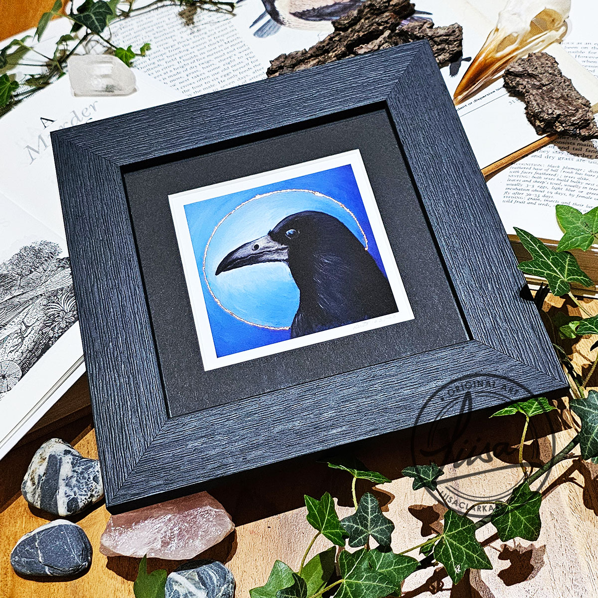 Angled photograph of a framed rook print with copper leaf detailing displayed with ivy and natural history books
