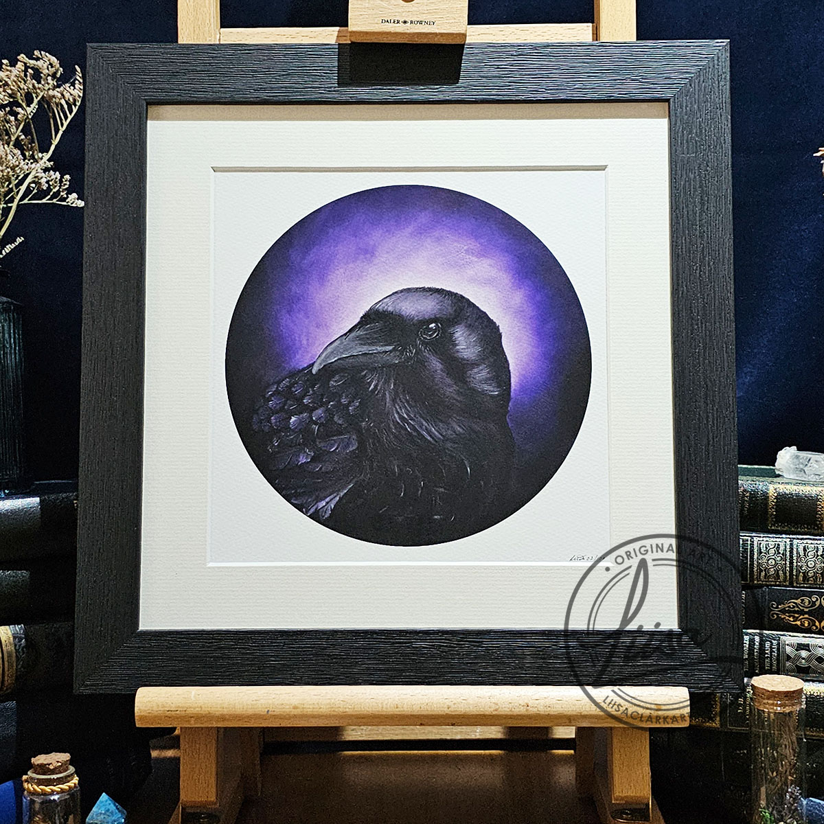 Close-up photograph of A Murder of Mauve by Liisa Clark Art, featuring a raven portrait with soft mauve and violet iridescent tones against a glowing purple circular background, displayed in a textured black frame on a wooden easel.