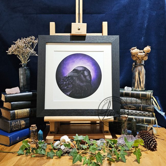 Framed raven artwork titled A Murder of Mauve displayed on a wooden easel surrounded by vintage books, ivy, crystals, dried seed heads and natural history inspired styling against a dark backdrop