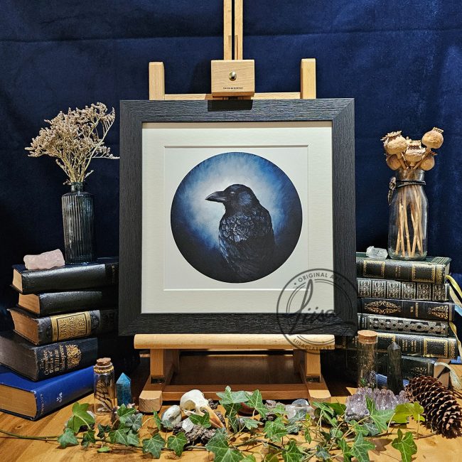 Framed raven artwork titled Prussian Blue displayed on a wooden easel surrounded by vintage books, ivy, crystals and natural history inspired objects against a dark backdrop.