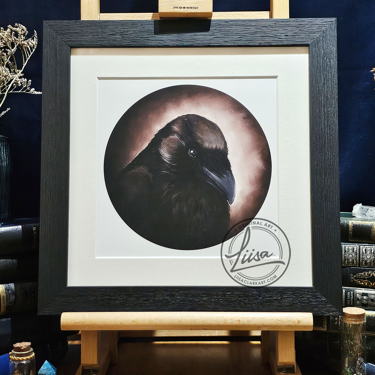 Close-up photograph of Sepia Storytelling by Liisa Clark Art, featuring a dark raven portrait surrounded by warm sepia and bronze tones, displayed in a textured black frame on a wooden easel.