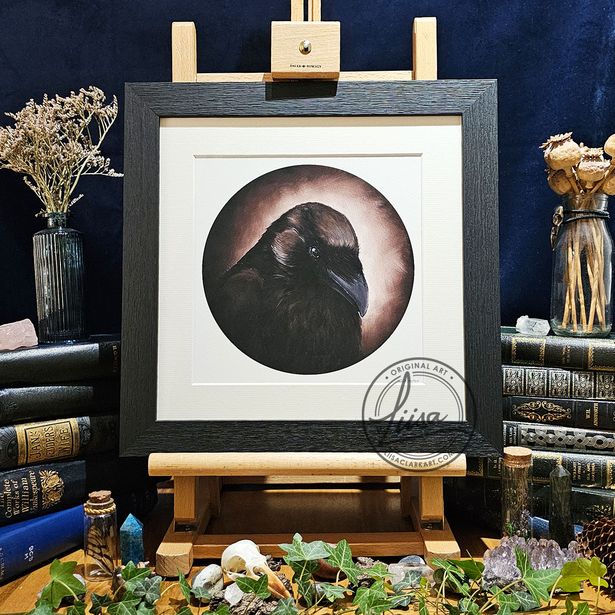Framed raven artwork titled Sepia Storytelling displayed on a wooden easel surrounded by vintage books, ivy, crystals and natural history inspired objects against a dark backdrop