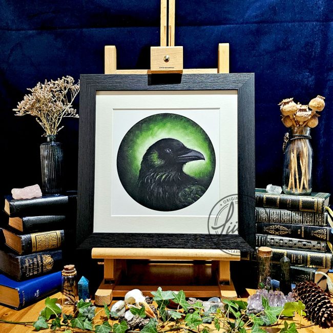 Framed raven artwork titled Verdant Green displayed on a wooden easel surrounded by vintage books, ivy, crystals, pinecones and natural history inspired styling against a dark backdrop