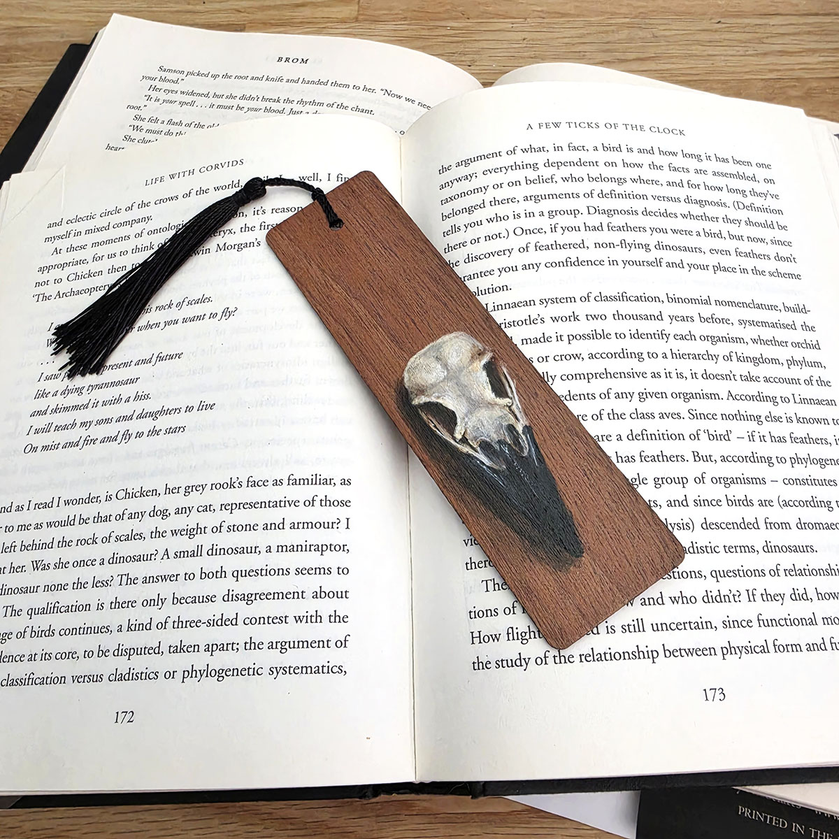 Hand-painted crow skull art bookmark on cherry wood with a black tassel, resting diagonally across the pages of an open book