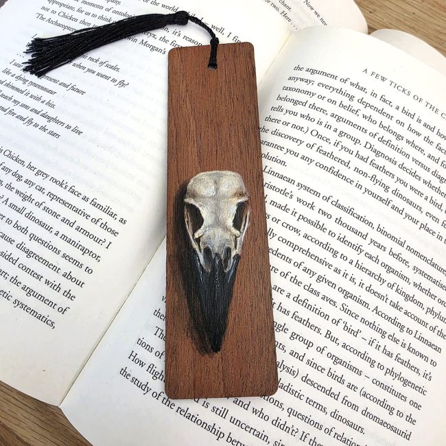 Original crow skull art bookmark hand-painted on cherry wood with a black tassel, displayed across the pages of an open book.