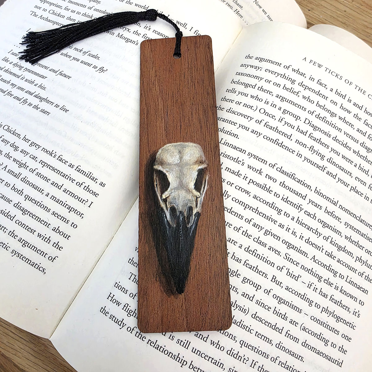 Original crow skull art bookmark hand-painted on cherry wood with a black tassel, displayed across the pages of an open book.