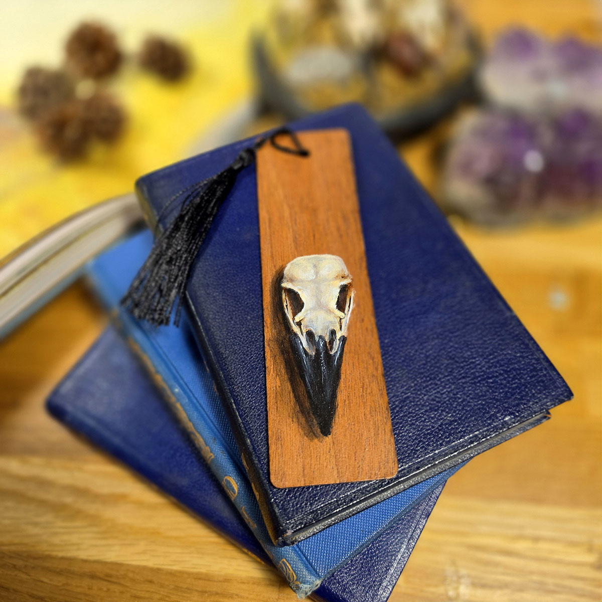 Hand-painted wooden bookmark featuring an original crow skull painting on cherry wood with a black tassel, resting on vintage books