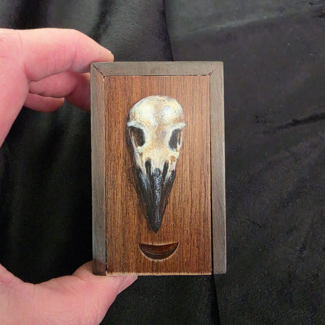 Handcrafted cherry wood trinket box with an original crow skull painting, held in the artist's hand to show scale.