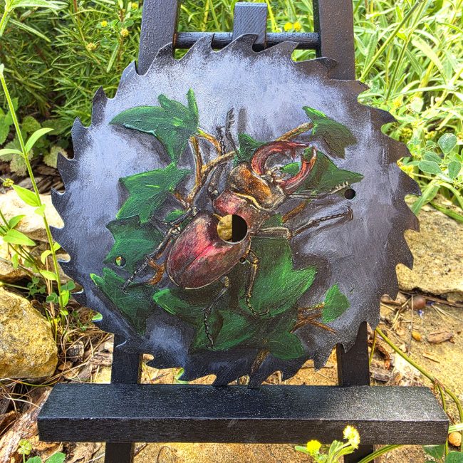 Original acrylic painting of a stag beetle on a reclaimed vintage circular saw blade displayed on an easel in a garden.