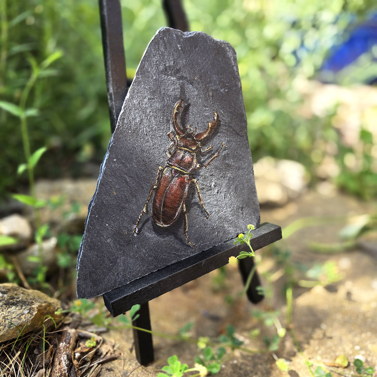 Keeper of the Old Stone original stag beetle painting displayed on an easel in a natural garden setting