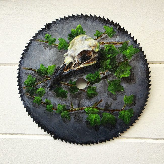 Original acrylic painting of a crow skull with ivy on a reclaimed vintage circular saw blade displayed on a white wall