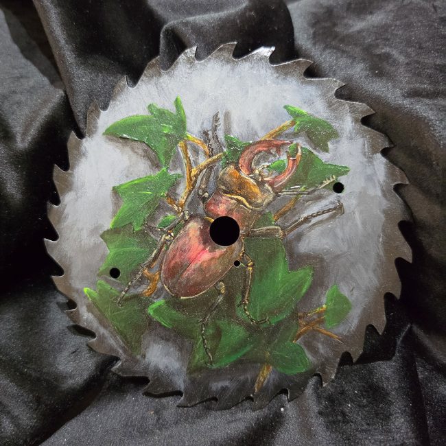 Original stag beetle and ivy painting on a reclaimed circular saw blade, photographed against black fabric