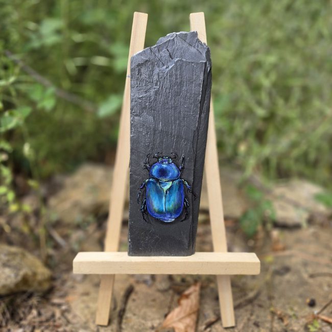 Blue beetle painting on reclaimed slate displayed on a small wooden easel outdoors
