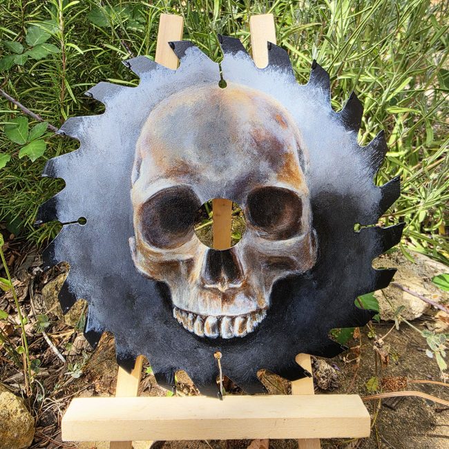 Close-up of Beneath Us All, a human skull painted on a reclaimed circular saw blade, displayed amongst garden foliage